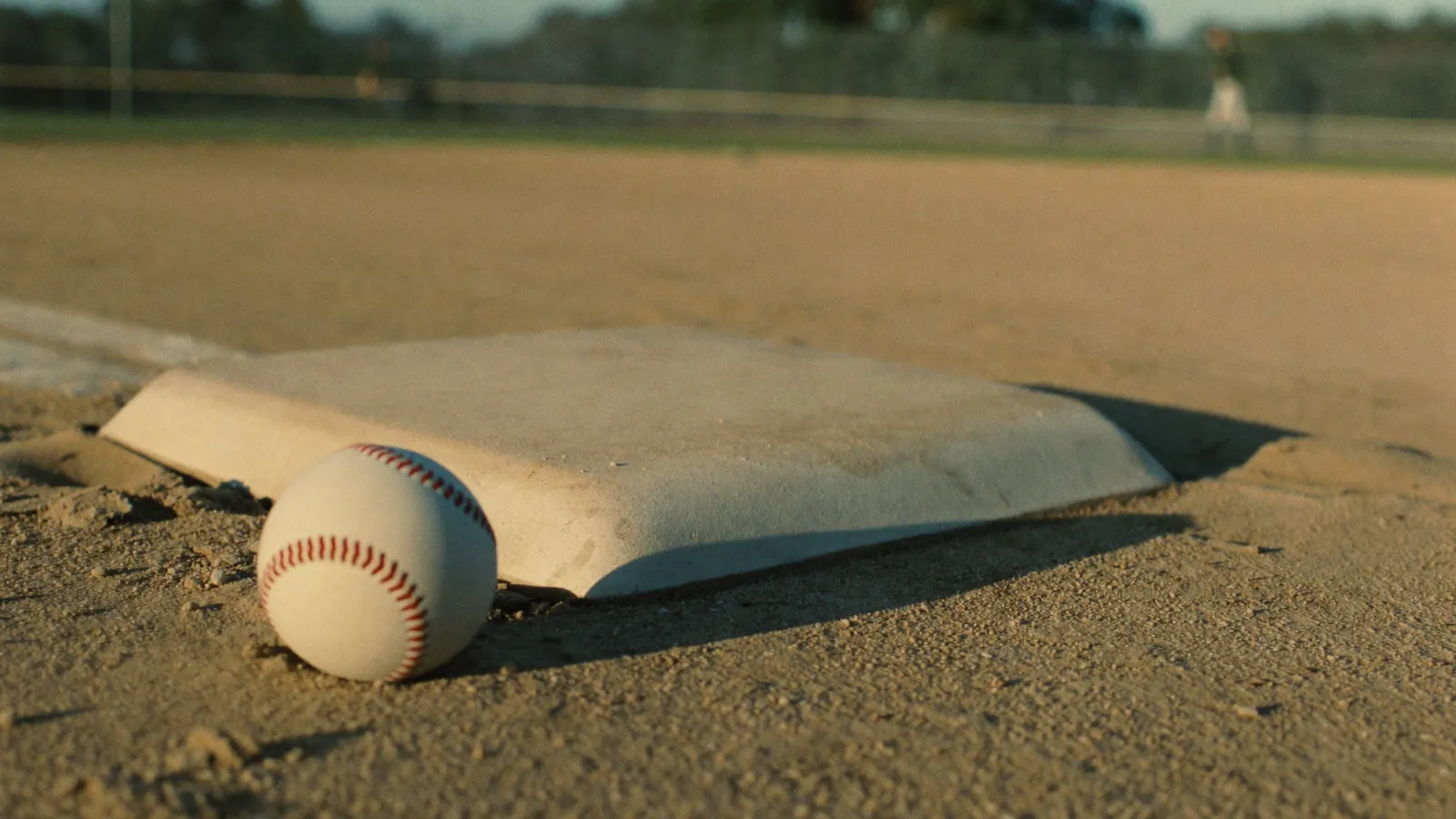 A baseball resting against a white base on the infield dirt at golden hour